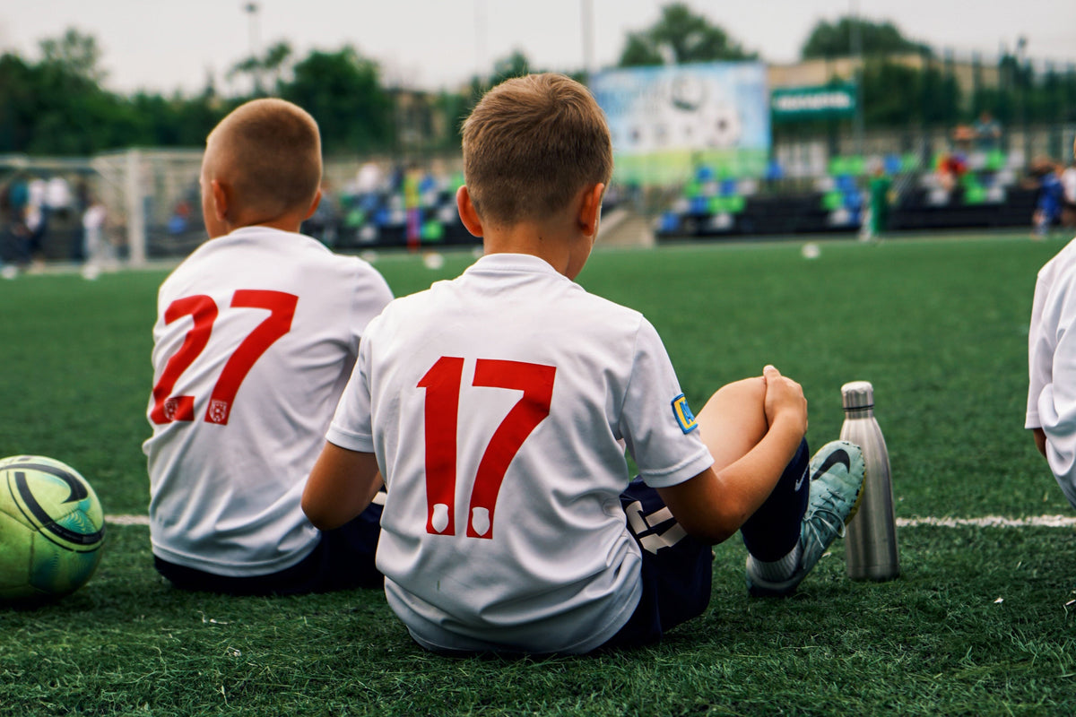 Boys in soccer jerseys sitting on a turf field during a summer game.