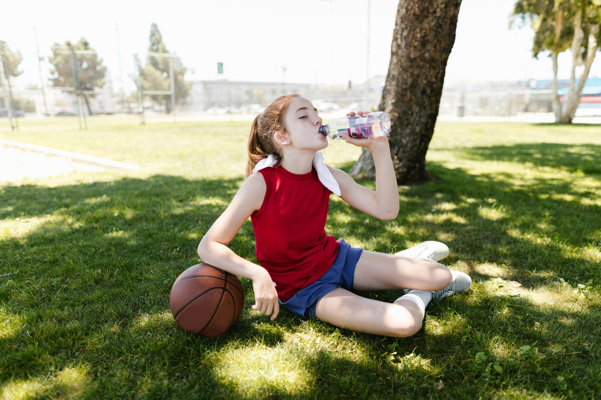 young girl in red tank top drinking water in a park with a basketball