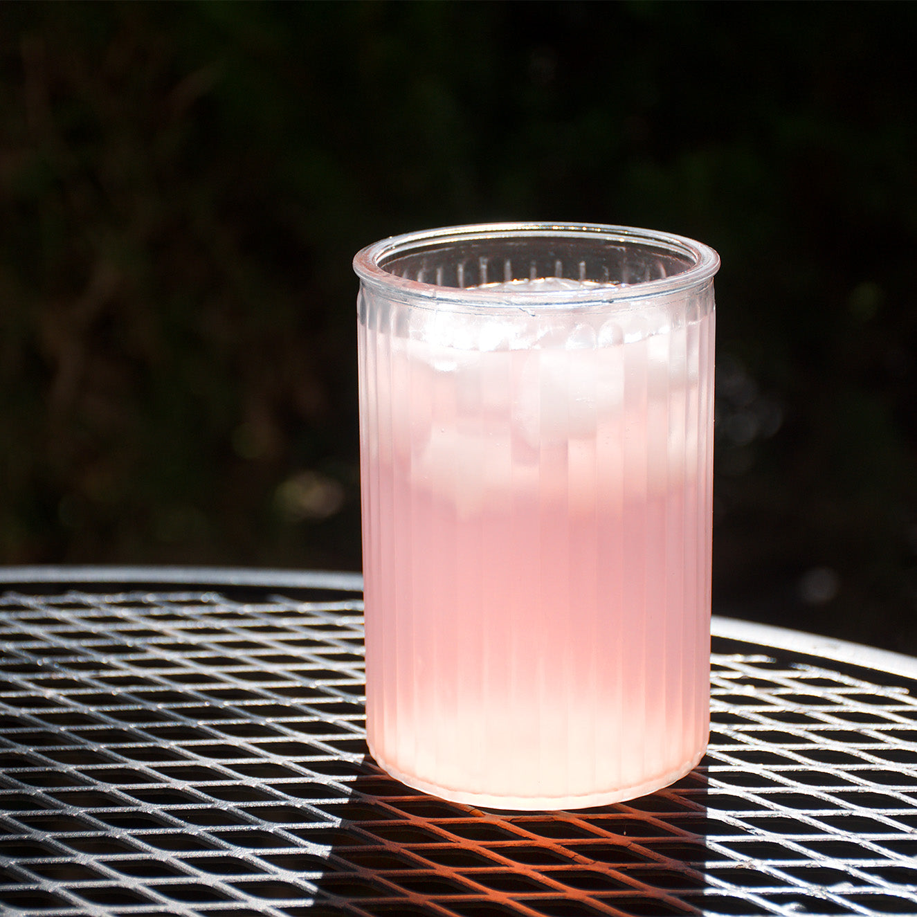 A clear ribbed plastic tumbler filled with a pink iced drink, placed on a black metal mesh table outdoors with a dark blurred background.